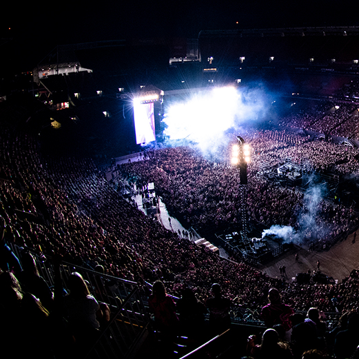 Stadium concert with packed crowd, intense lighting and smoke